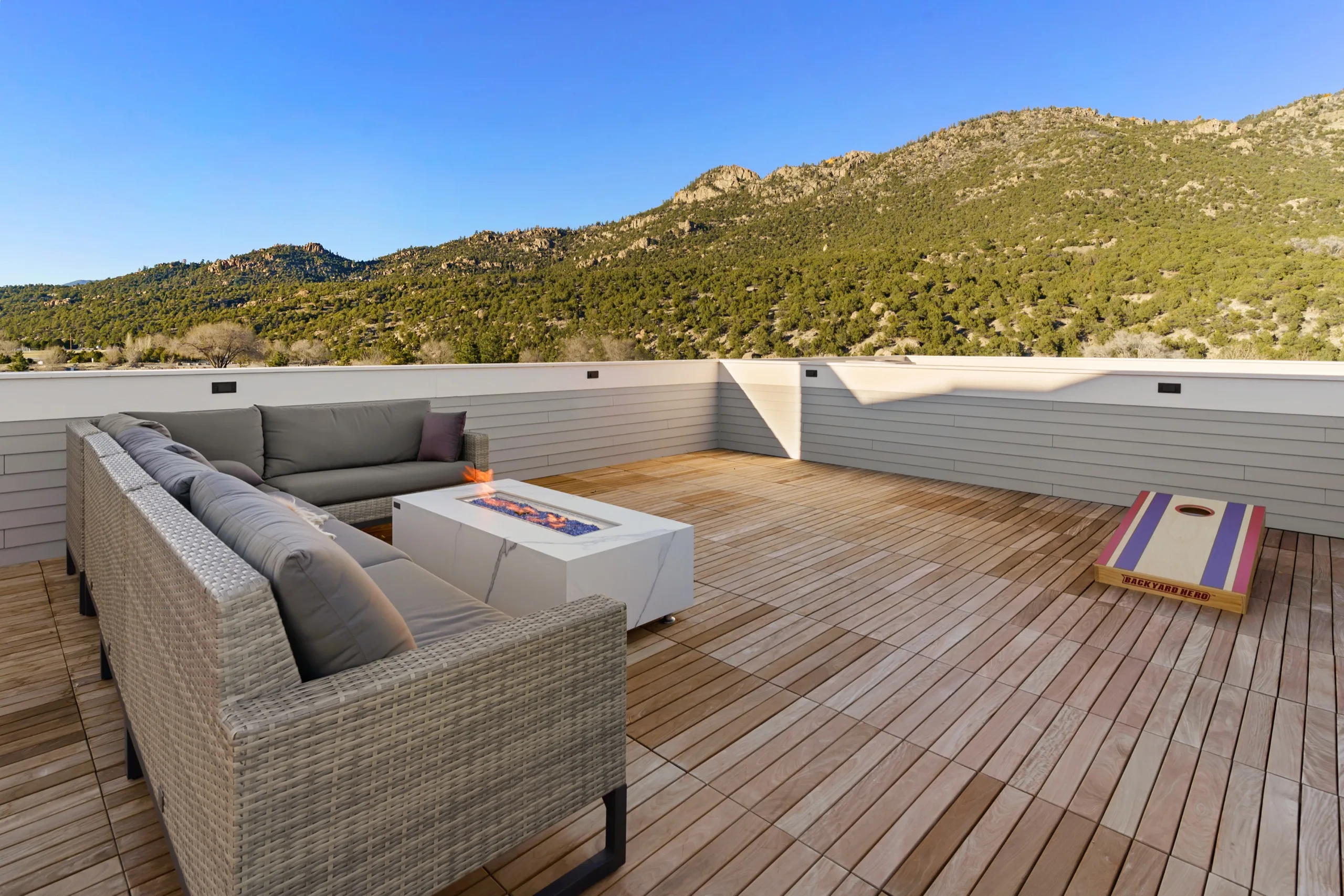 Rooftop deck with a wicker sectional drawn round a fire table, the mountain ridge filling the view past the parapet