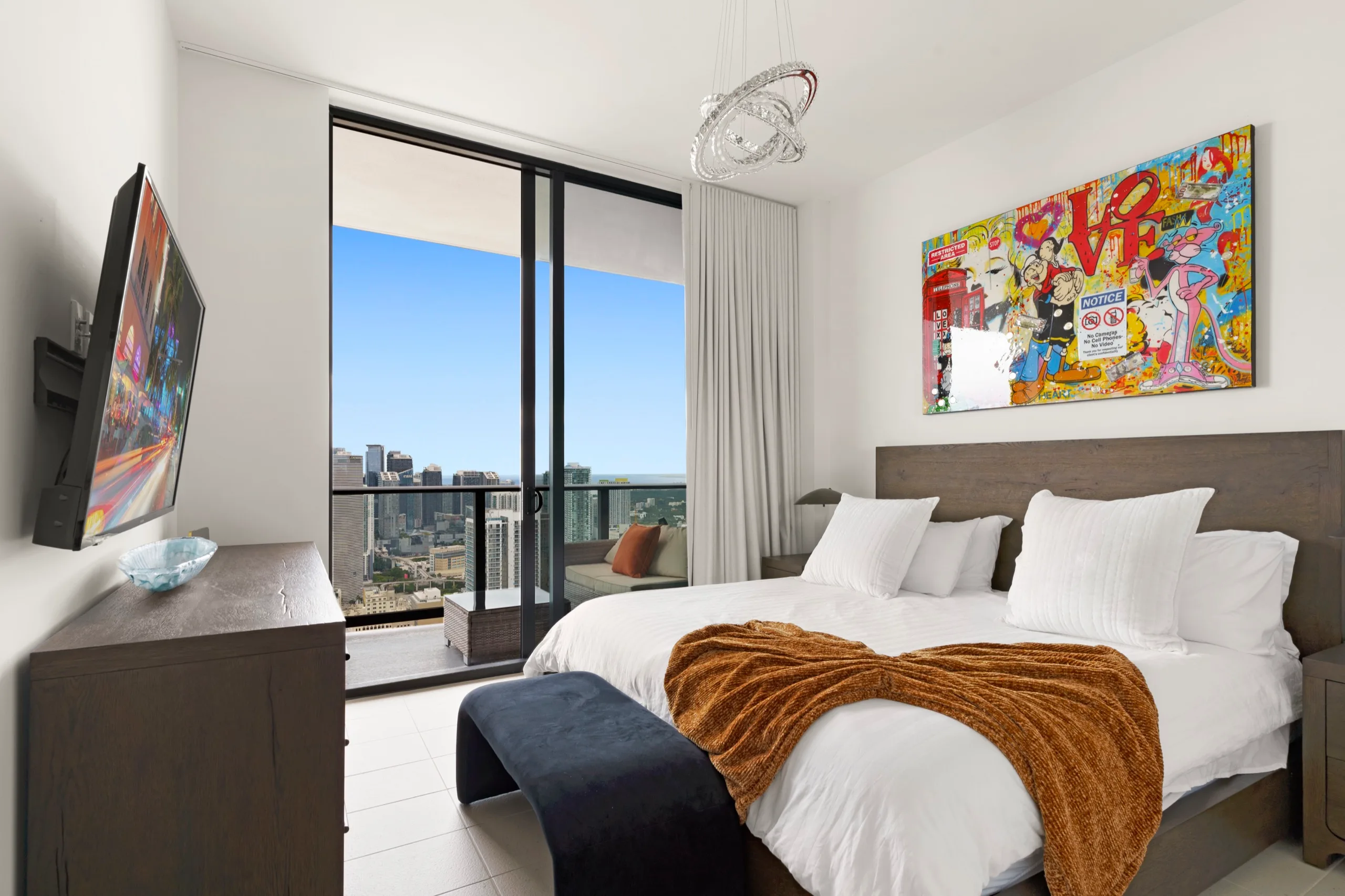 High-rise bedroom with a caramel throw and a pop-art canvas, floor-to-ceiling glass onto the Miami skyline