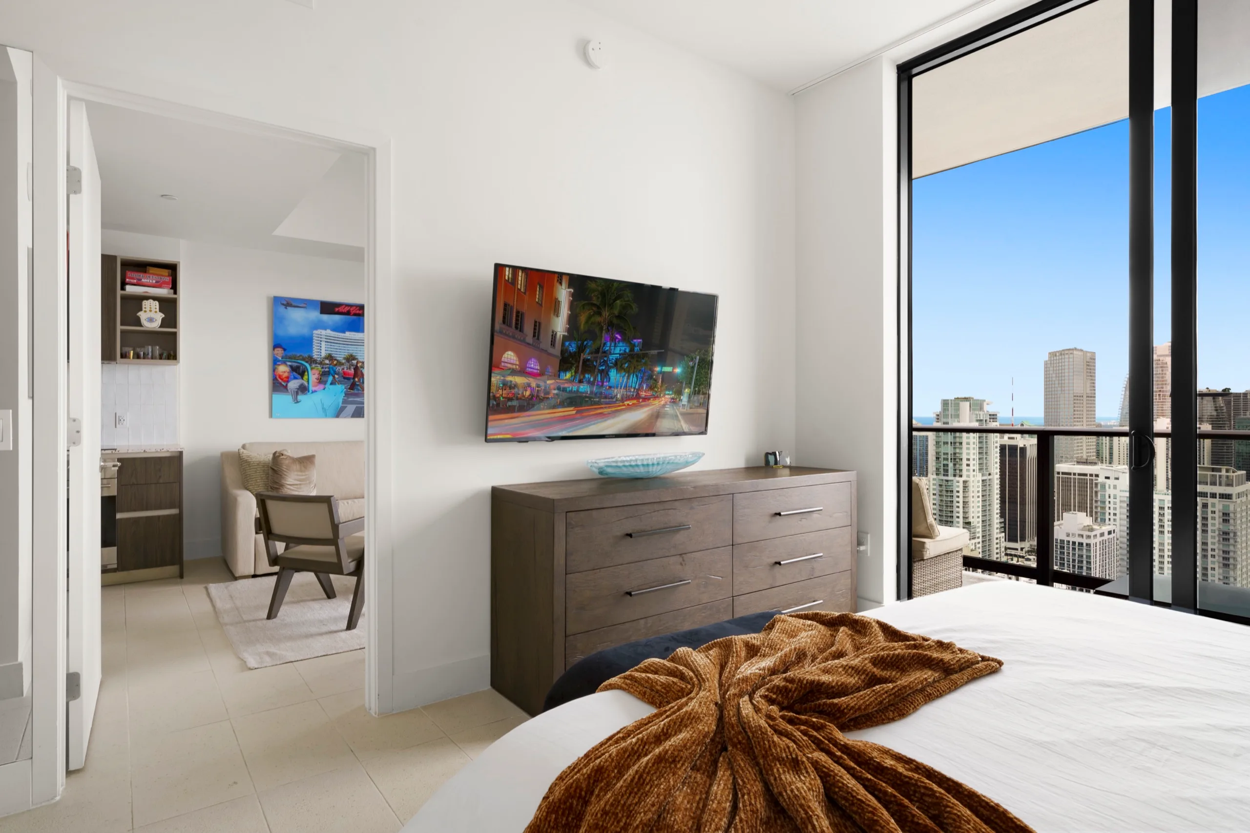 Bedroom dresser and wall-mounted screen beside a balcony door, downtown towers beyond the glass