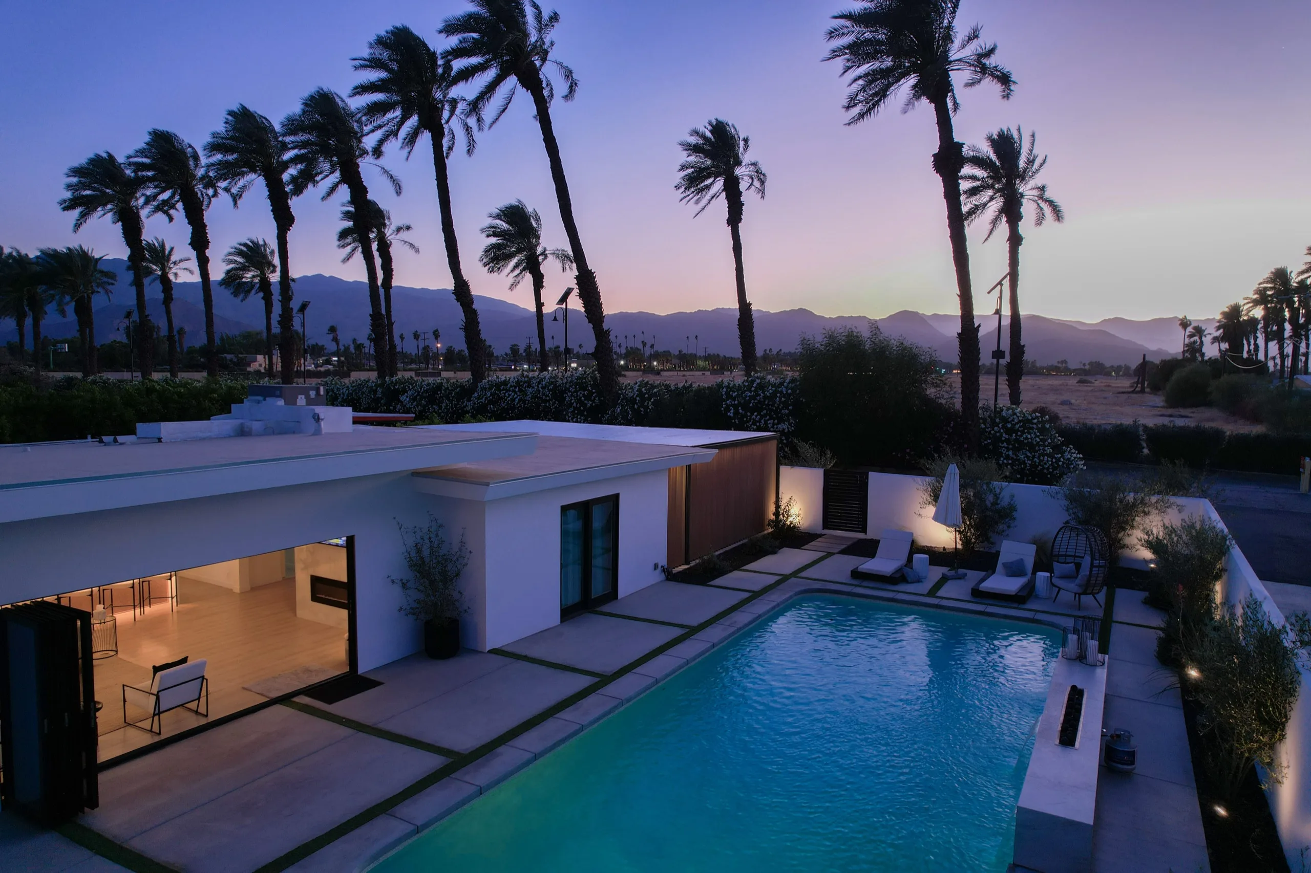 Aerial view at dusk of a walled desert house, lit pool and palm court glowing against the mountains