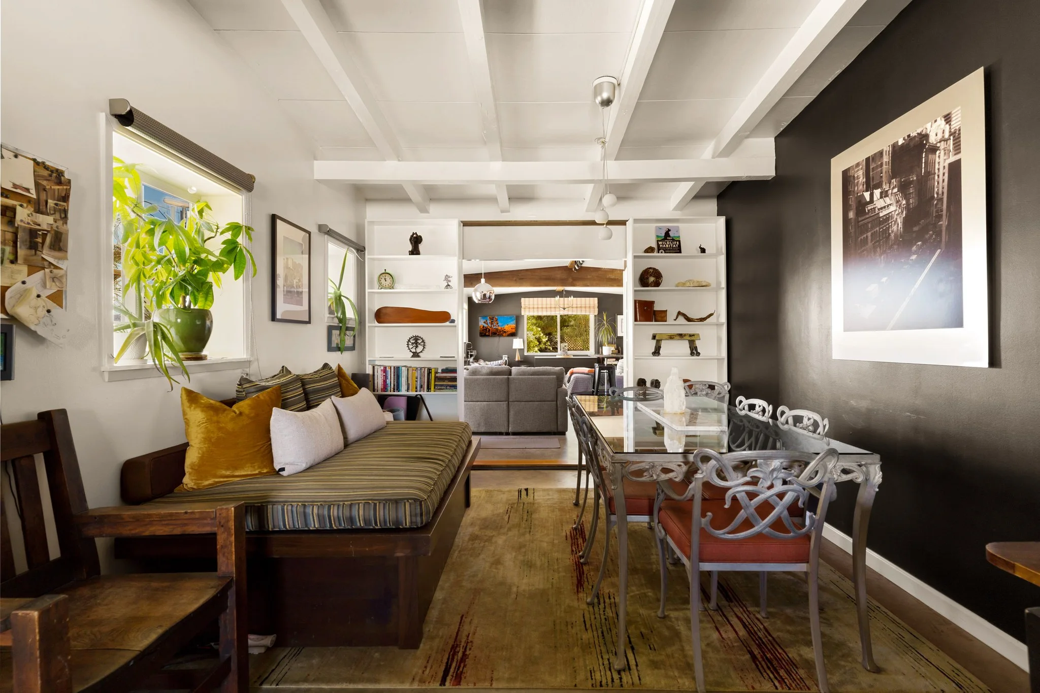 Living and dining room under white-painted beams, a black gallery wall of prints and a glass table ringed with sculptural chairs