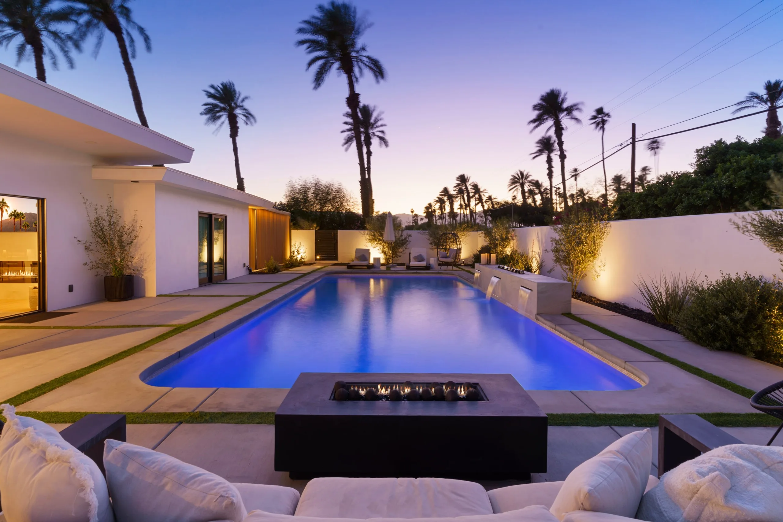Desert pool at dusk with a lit fire table in the foreground, palms silhouetted against a violet sky