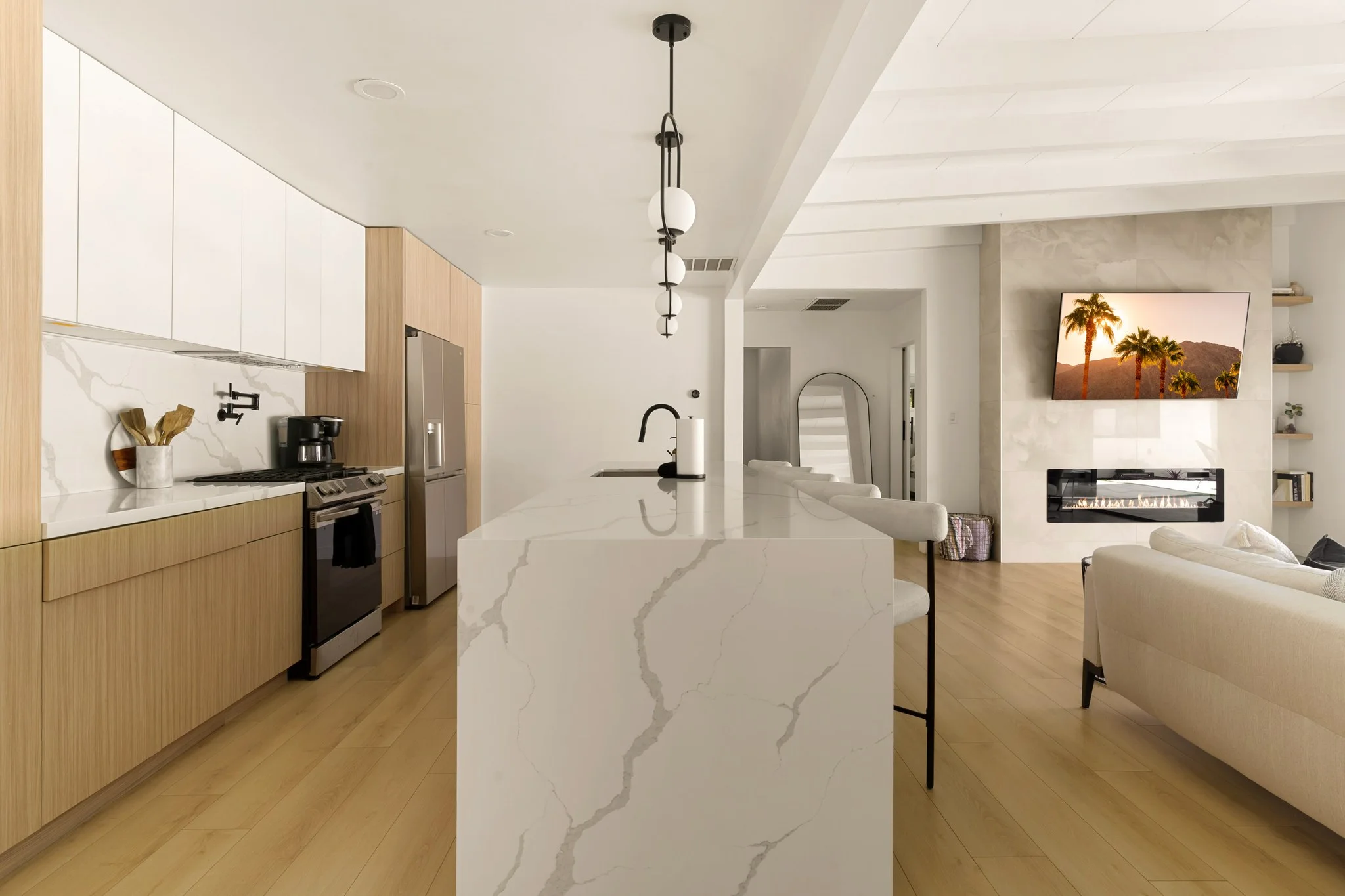 Kitchen island in white marble under exposed beams, pale oak cabinets and the living room beyond