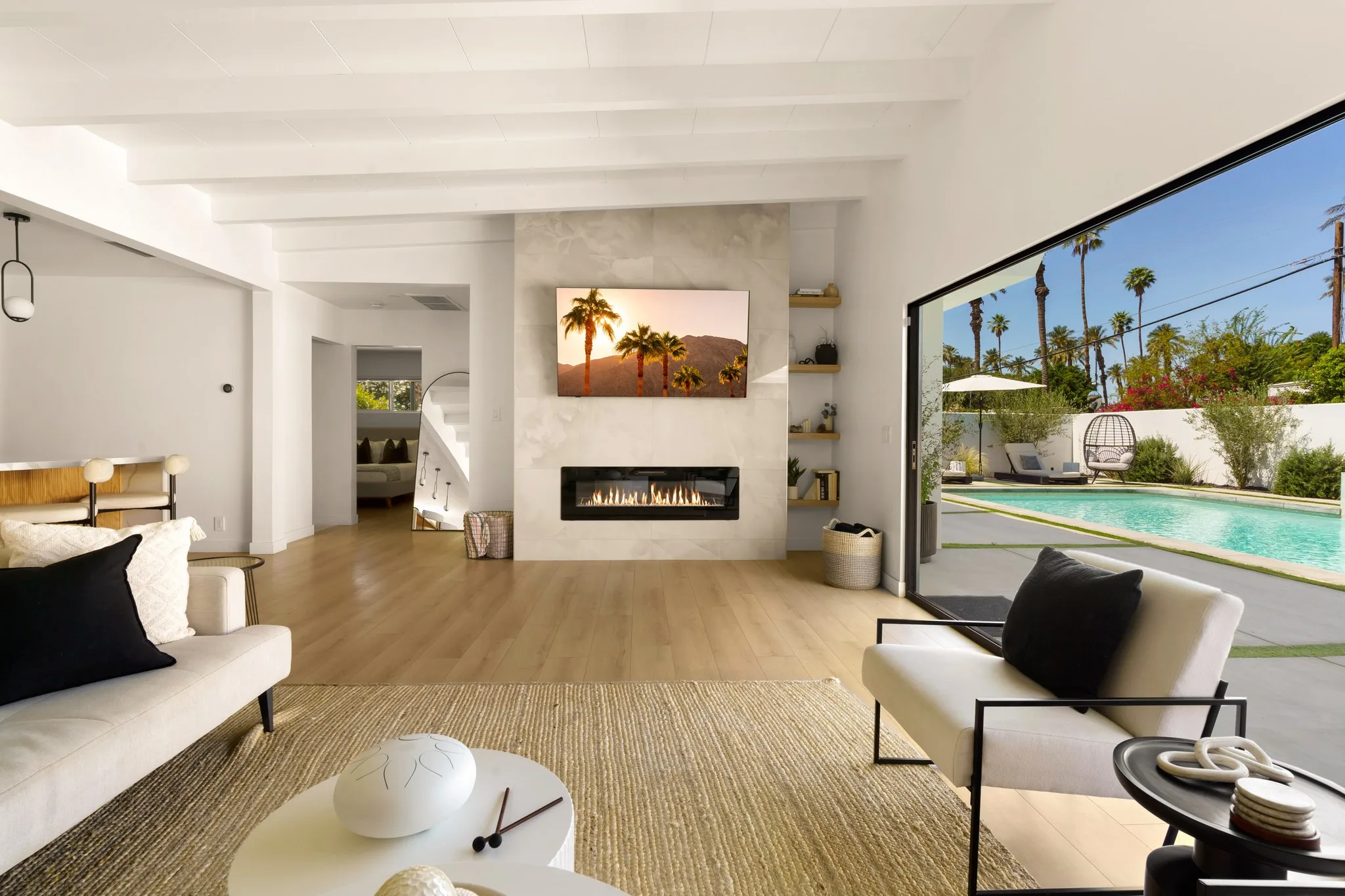 Living room in white and pale oak, linear fireplace below a desert photograph, glass wall onto the pool