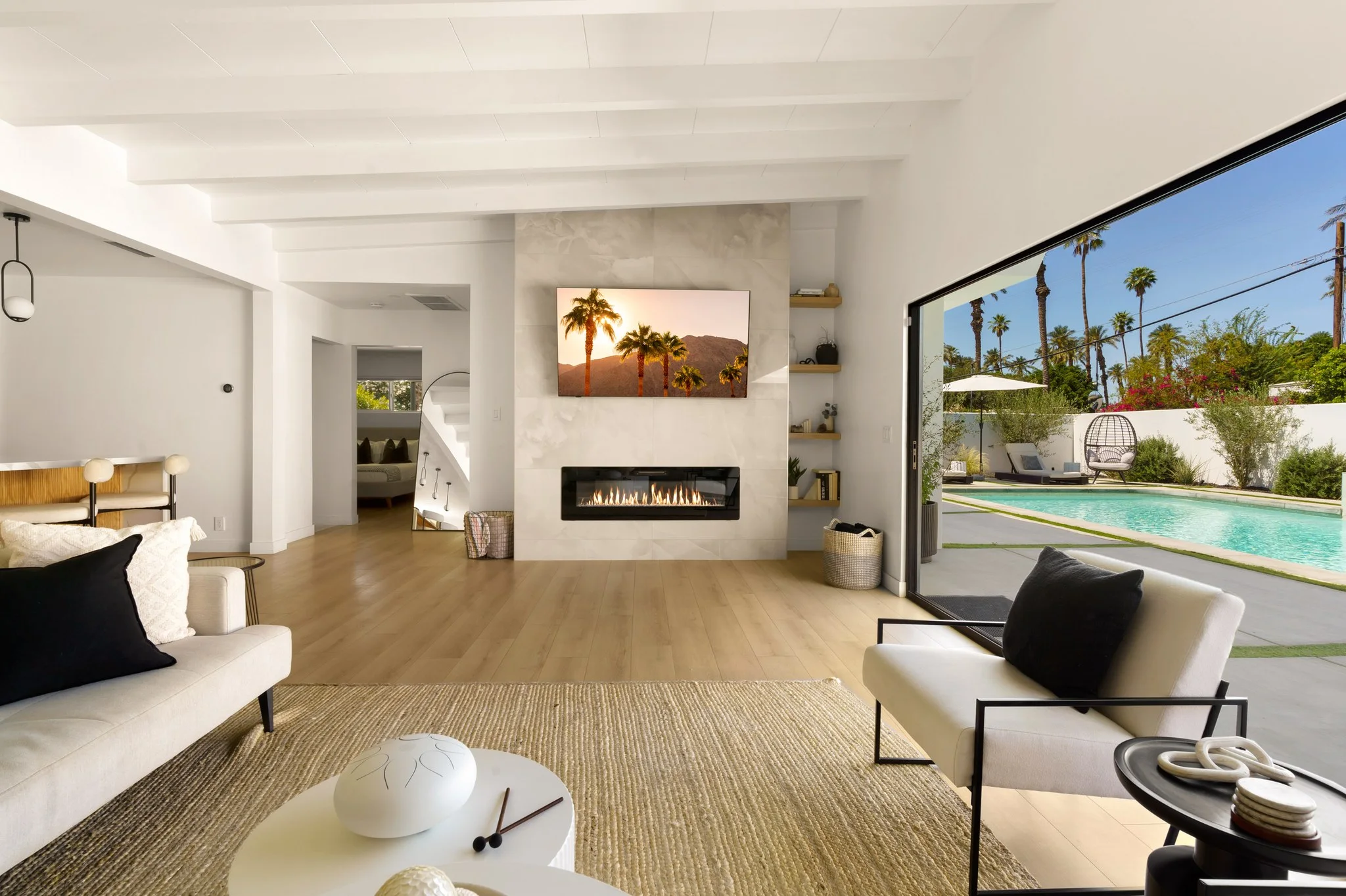 Living room in white and pale oak, linear fireplace below a desert photograph, glass wall onto the pool