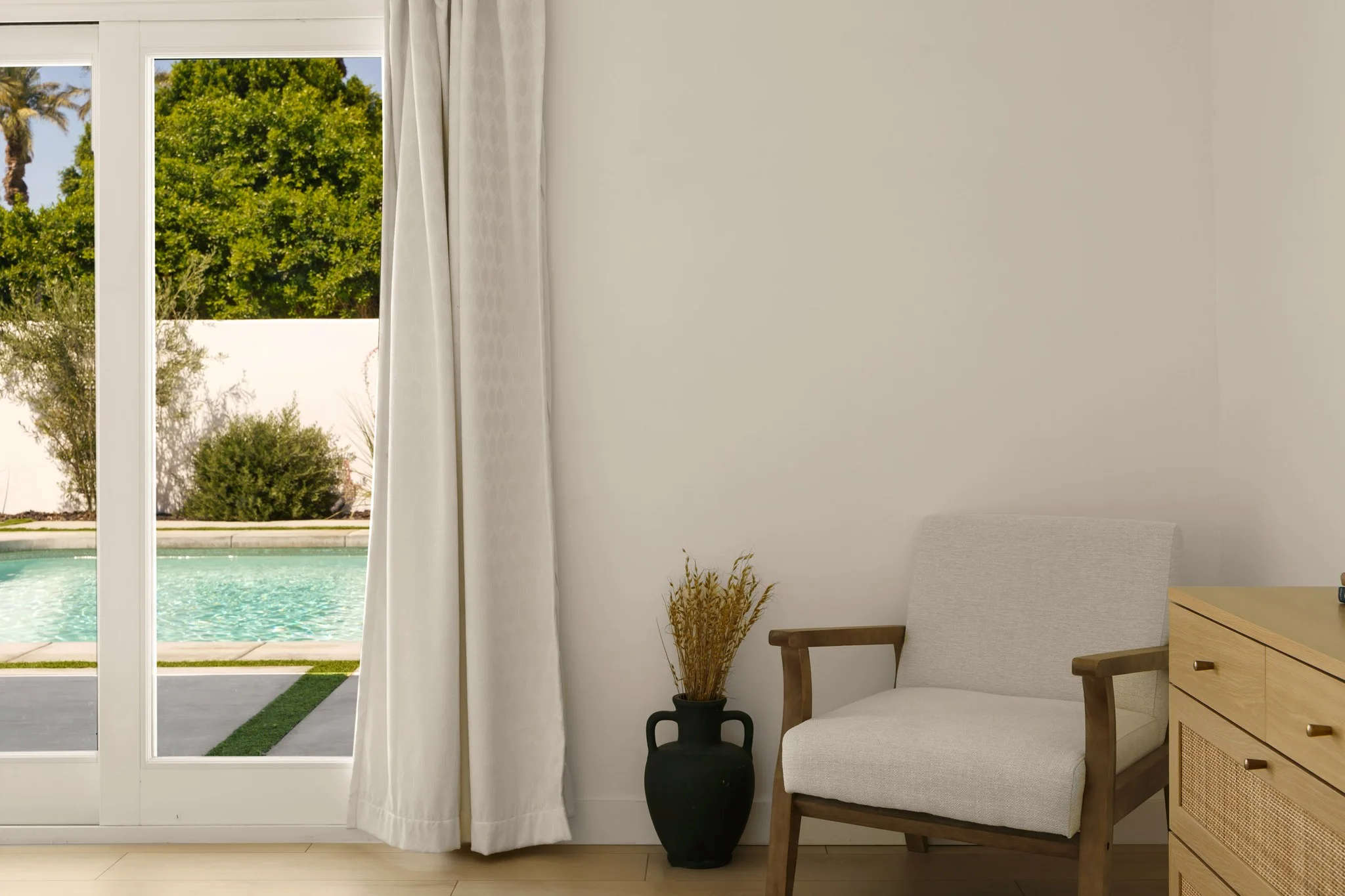 Cream armchair and oak dresser beside a window onto the pool, dried grasses in a black urn