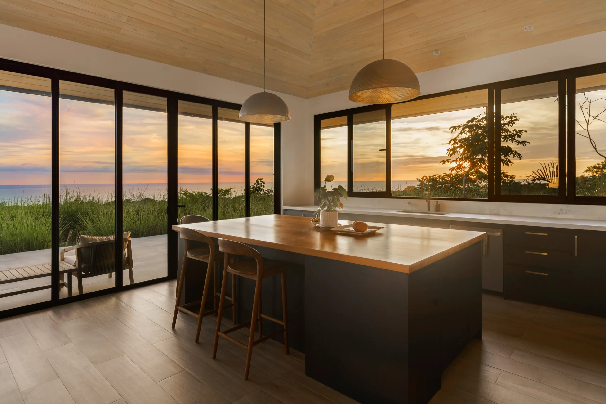 Kitchen island under a timber ceiling at sunset, sliding doors open to a deck above the ocean