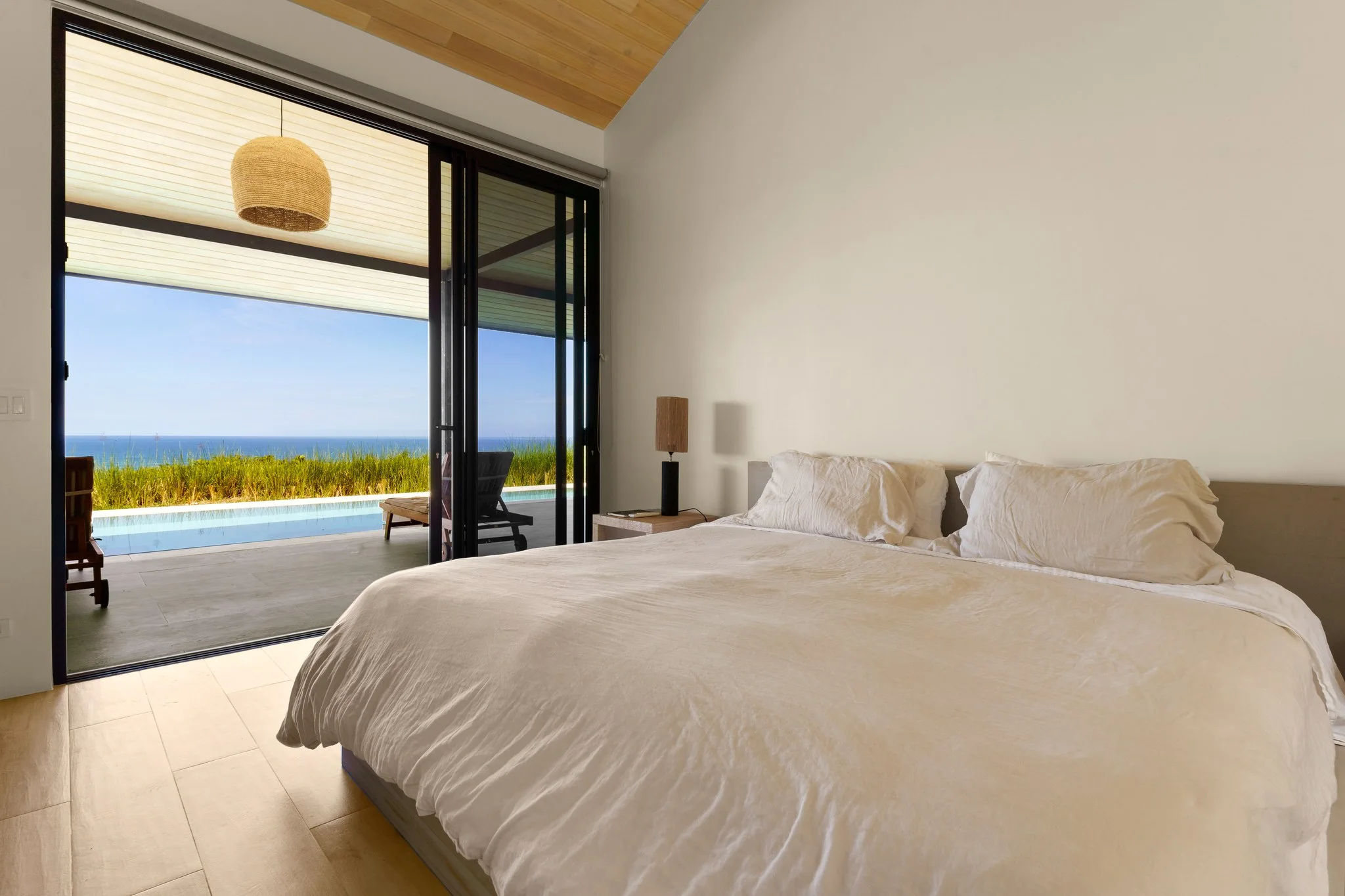 Bed made in white linen facing a wall of glass folded open to the pool and the ocean horizon
