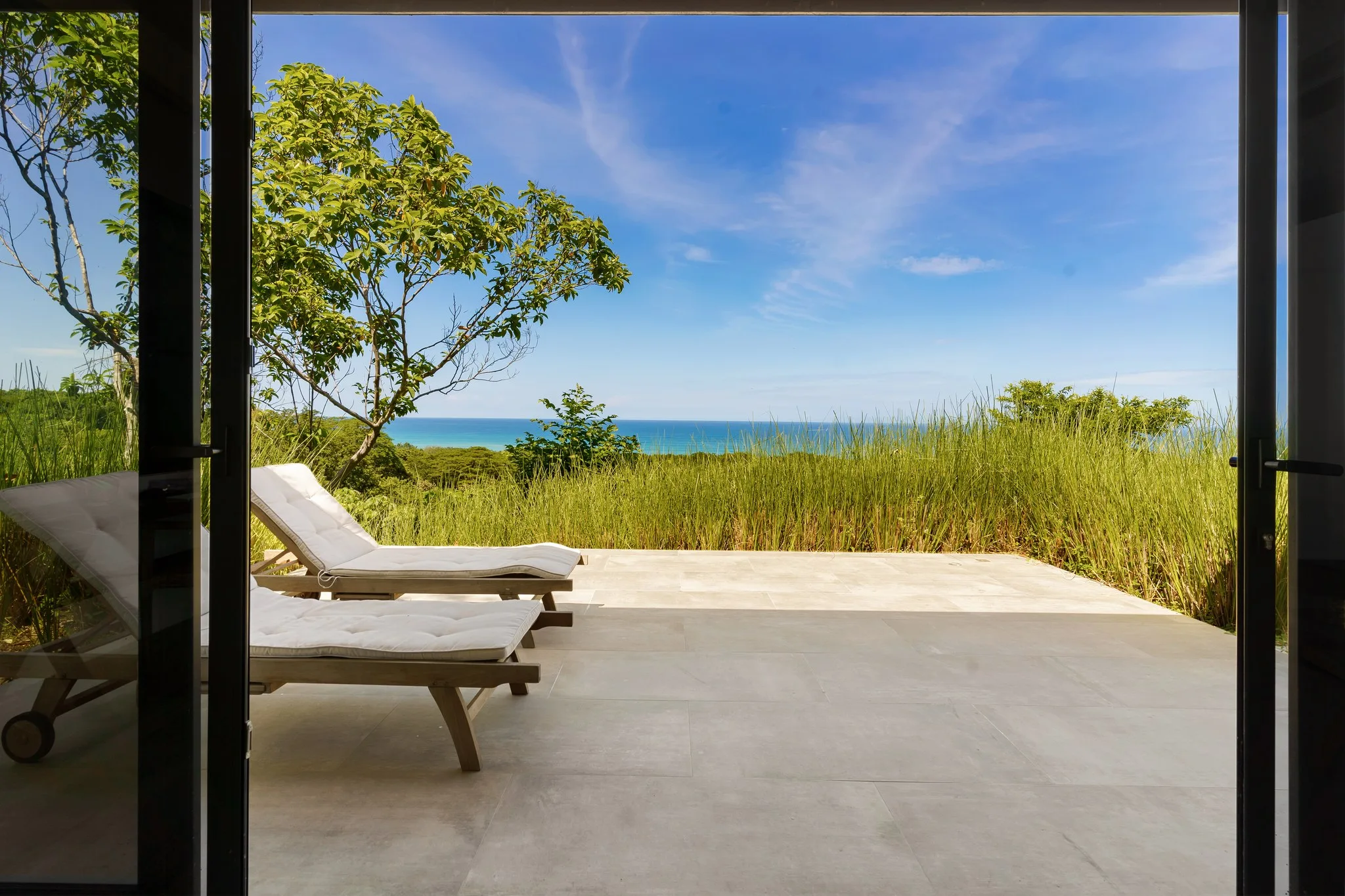 Loungers on the terrace at the edge of the dune grass, the sea flat and blue beyond