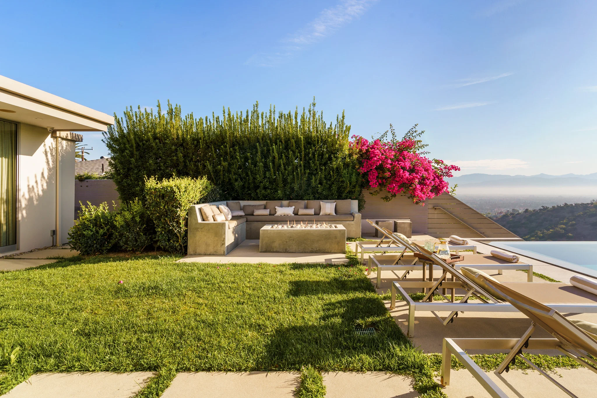 Clipped lawn running to a pool terrace and outdoor kitchen, hedge and flowering vine screening the view beyond