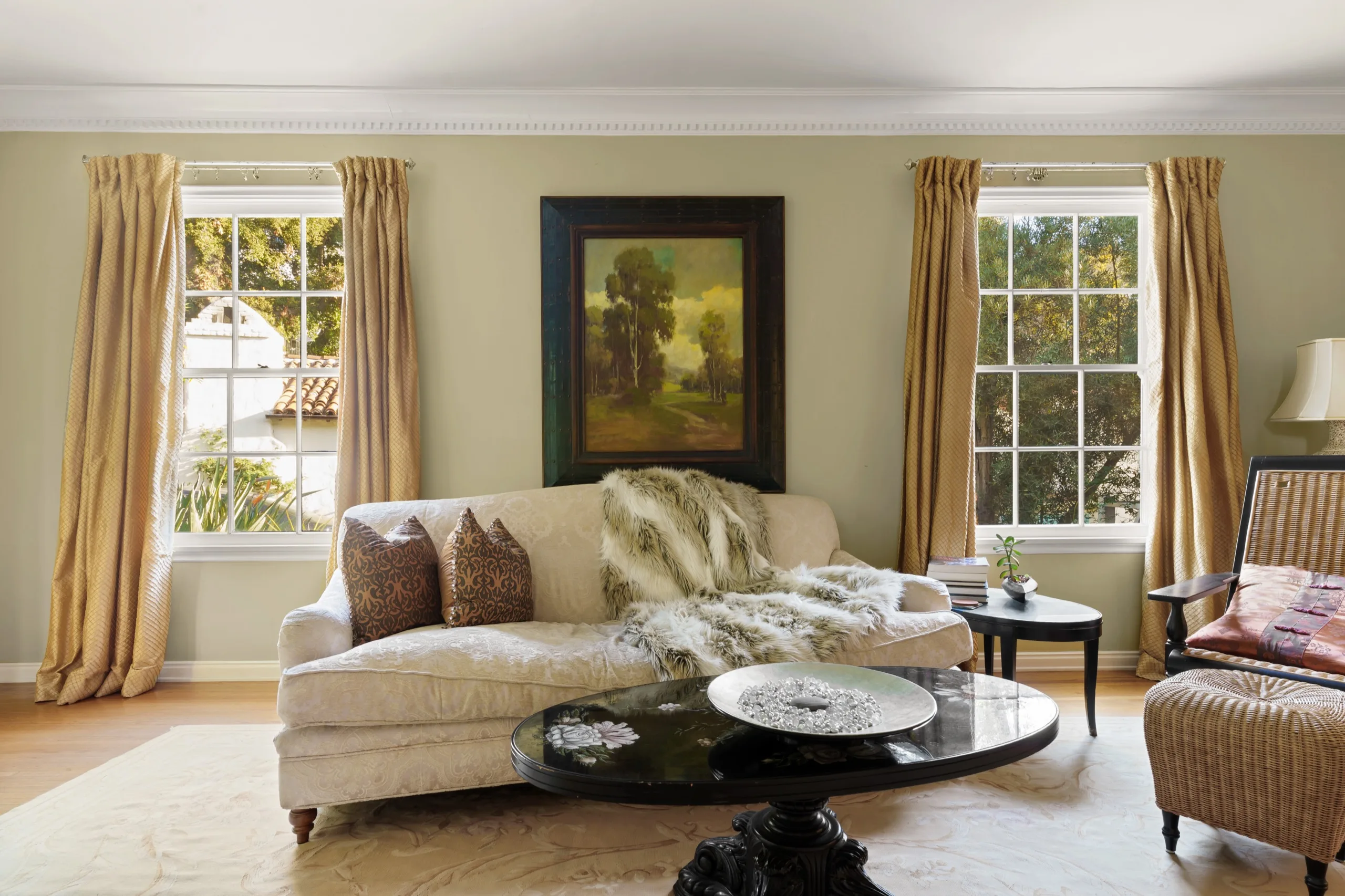 Living room in gold and cream, tall curtained windows either side of a framed landscape above the sofa