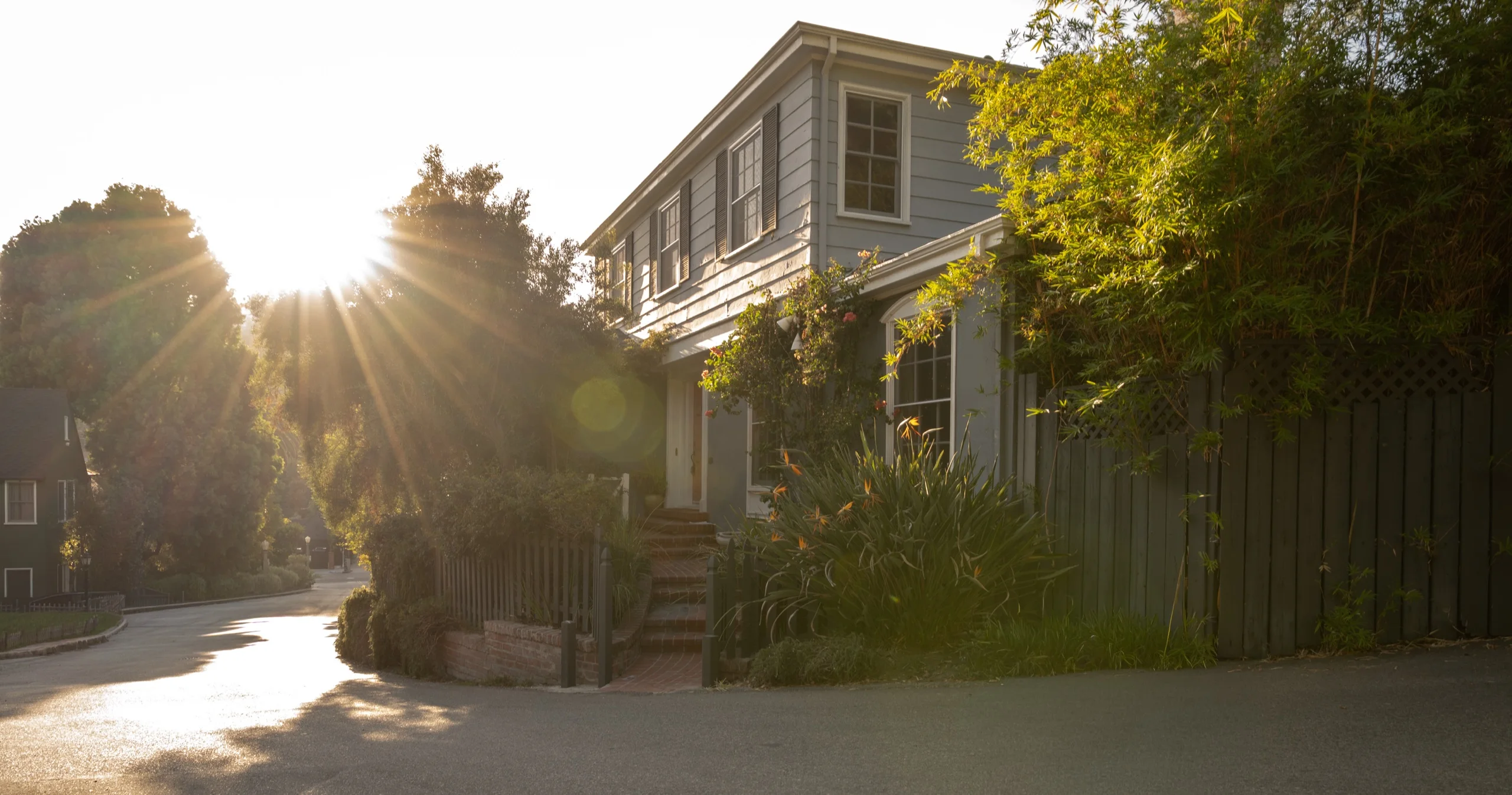 The house at golden hour, sun breaking through the trees across a clapboard facade and a planted front path