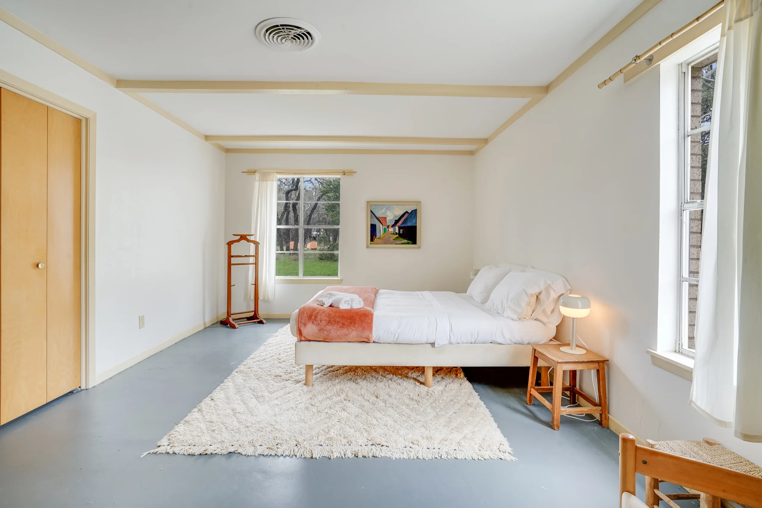 Bare, light-filled bedroom with a low timber bed, sheepskin rug and tall shuttered windows