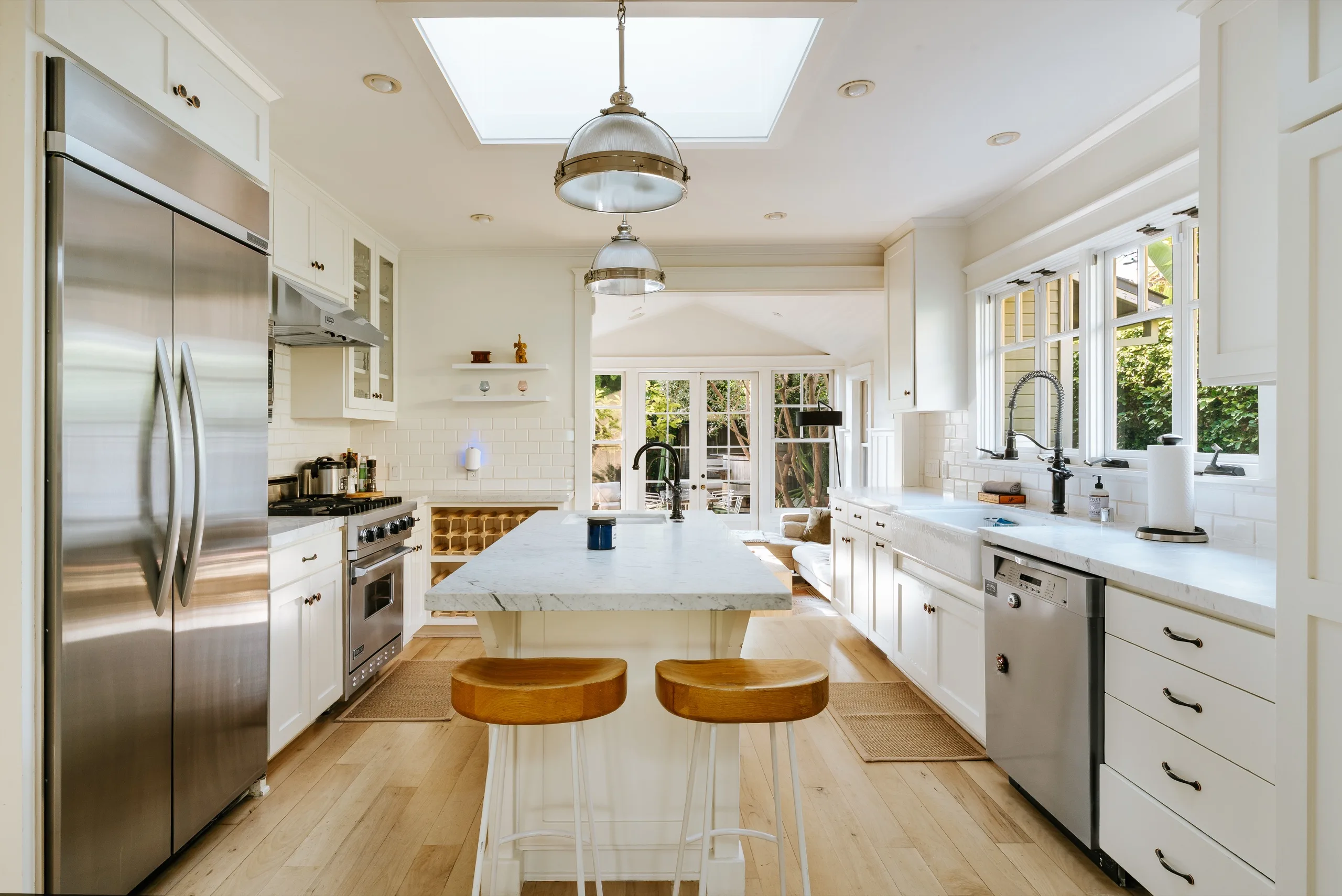 White kitchen beneath a skylight, marble island with leather counter stools and a run of garden doors