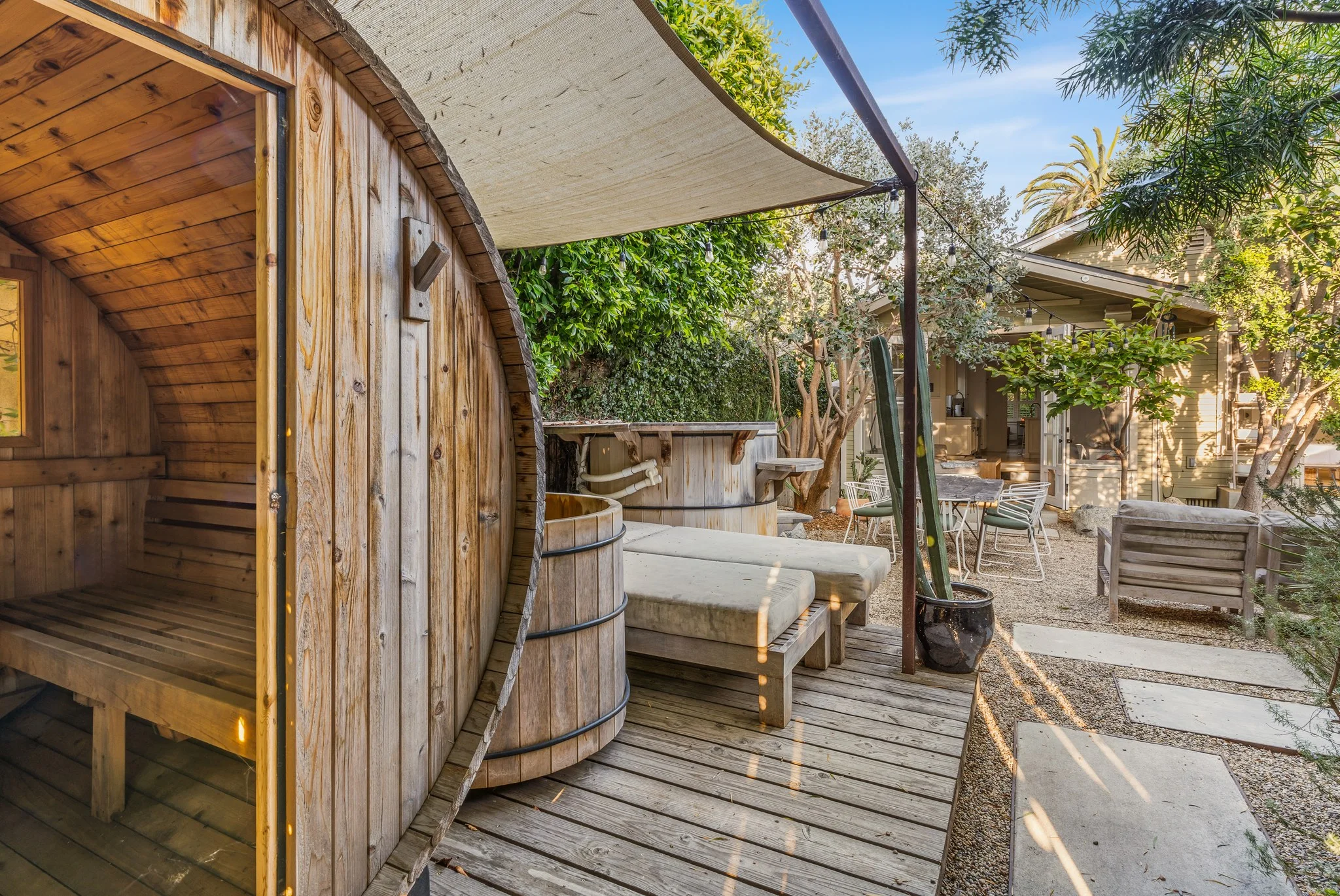 Barrel sauna and a timber hot tub on a deck under a shade sail, palms and planting beyond