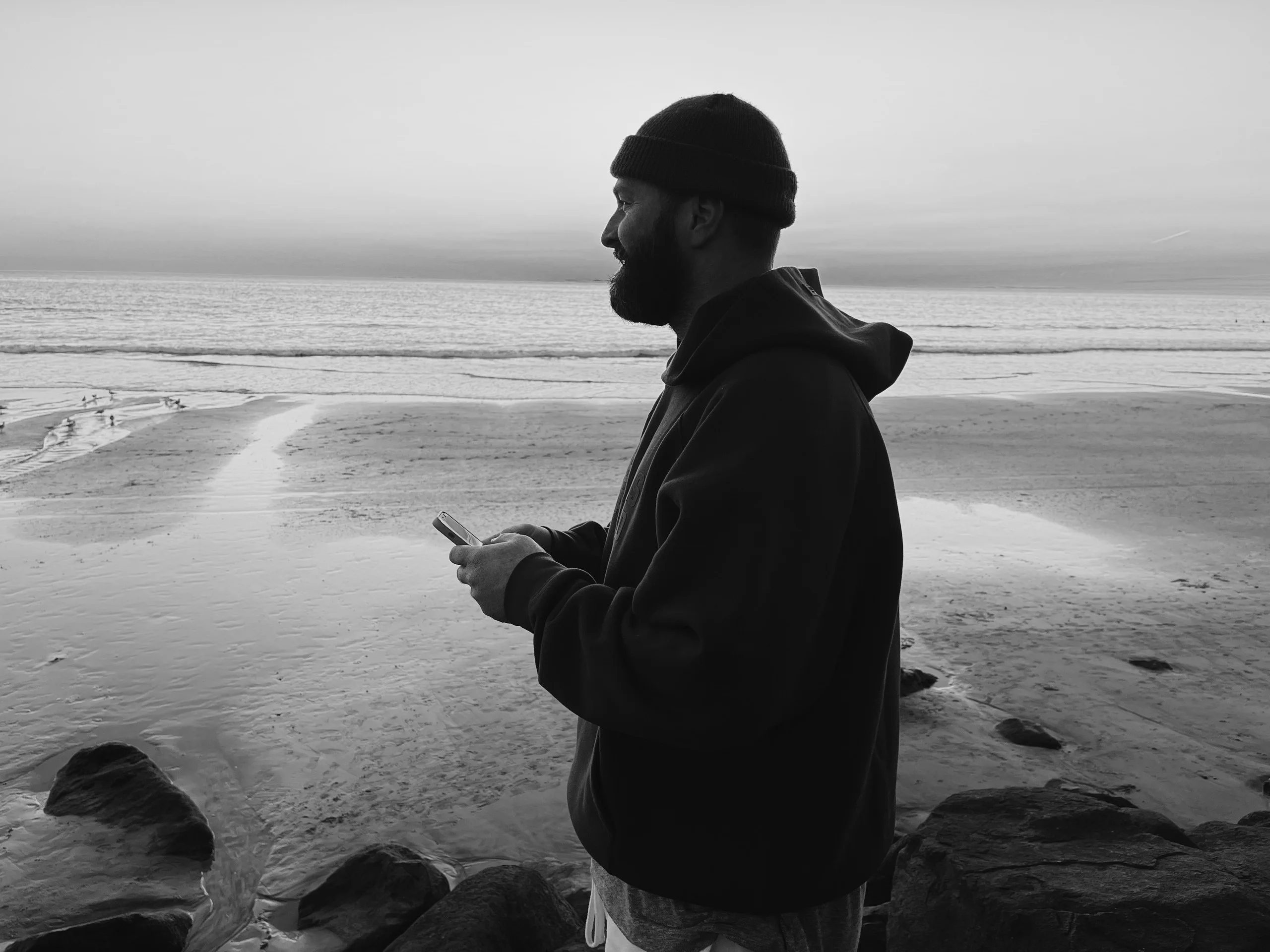Evan Haskell on an empty beach at first light, phone in hand, looking out over the wet sand to the water