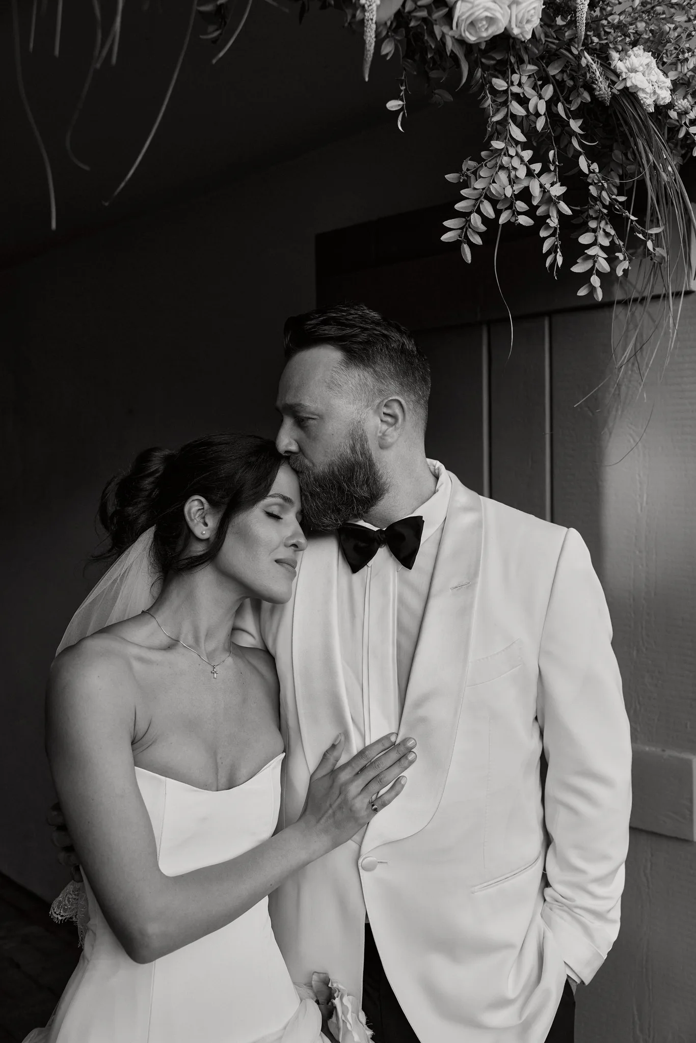 Evan and Dakota on their wedding day in black and white, he in a white dinner jacket kissing her forehead, flowers overhead
