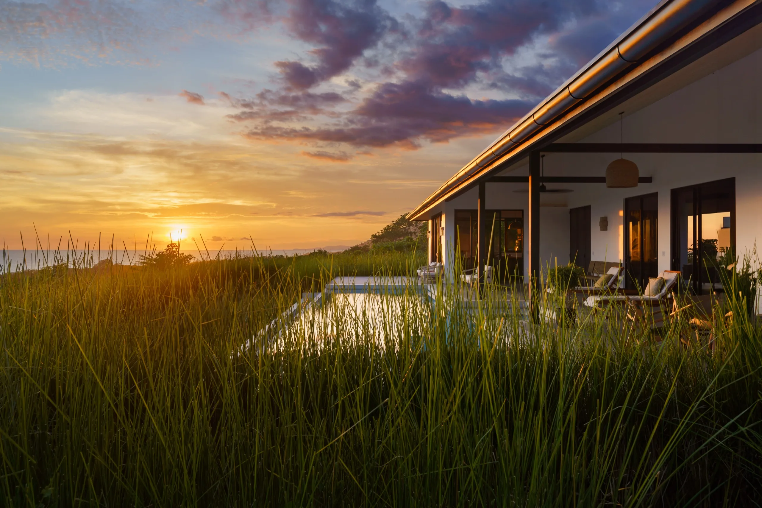 Coastal house at sunset seen across dune grass, lap pool catching violet and gold cloud light