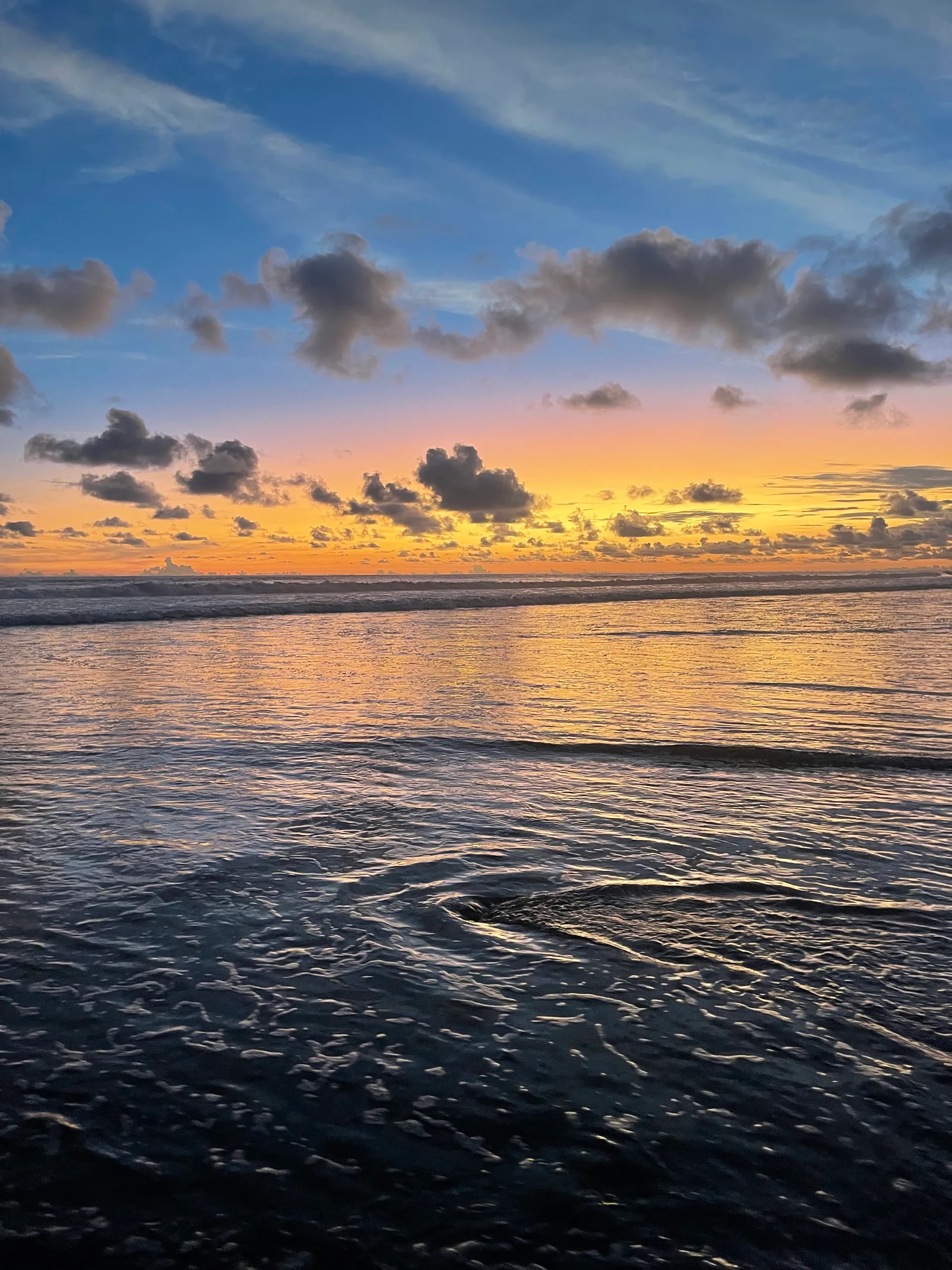 Wet sand and a slow eddy of water reflecting a pink and gold sunset sky