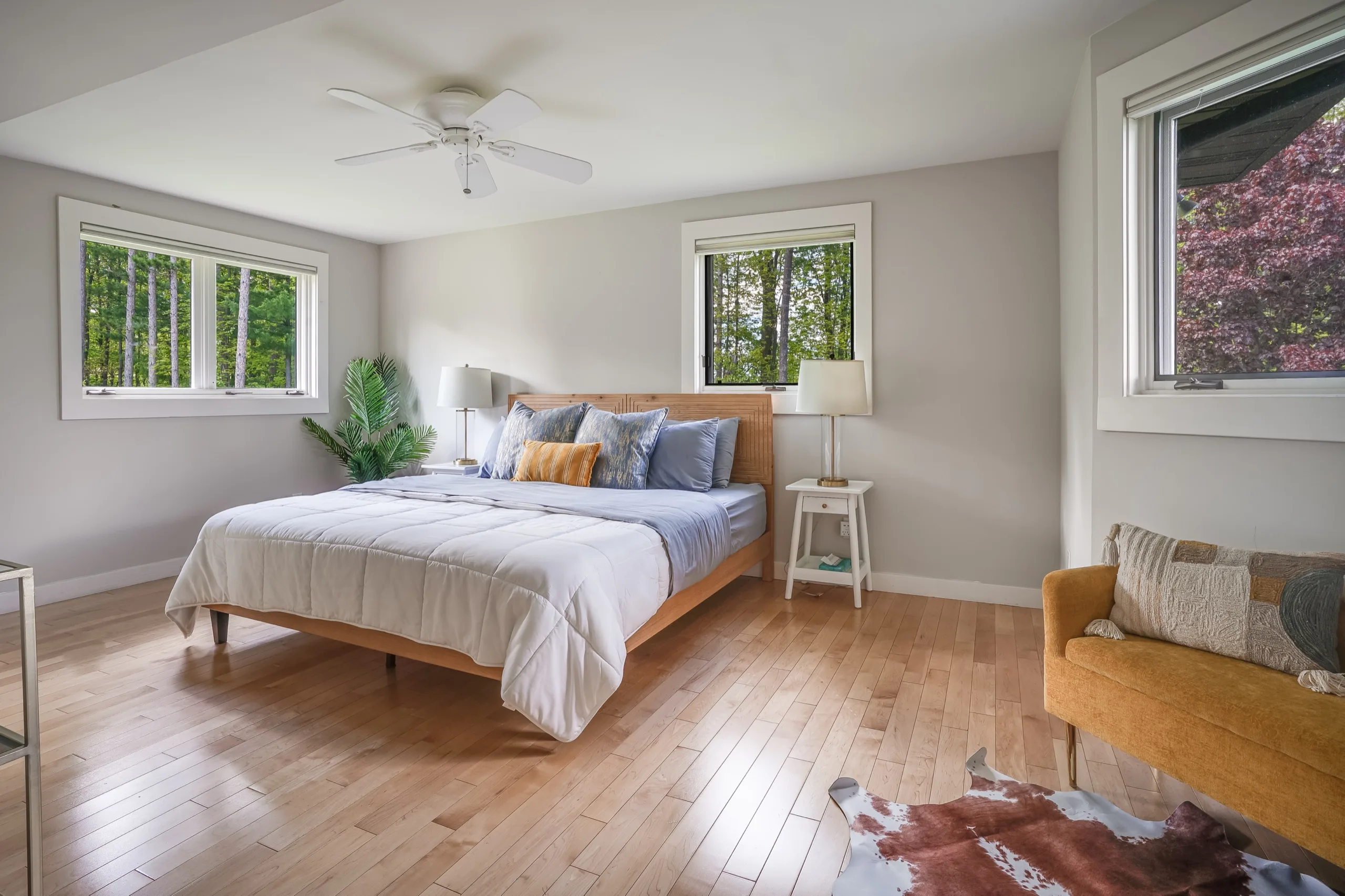 Bedroom in soft morning light, twin windows onto the trees and a cowhide rug across pale oak boards