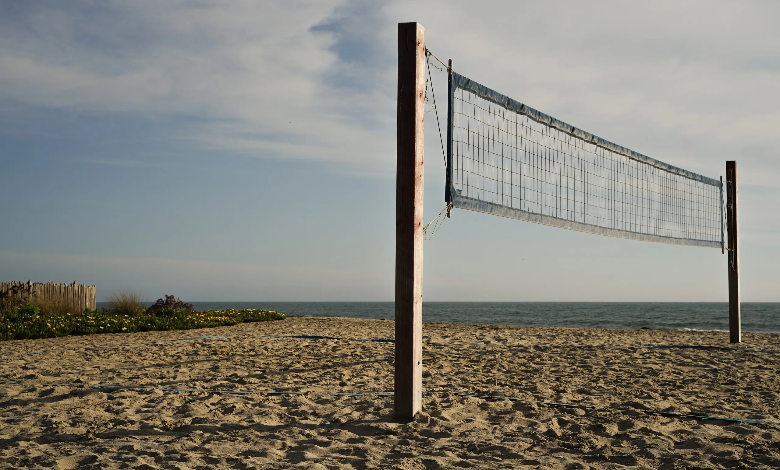 Beach volleyball net strung between weathered posts on empty sand, the ocean and a pale sky behind